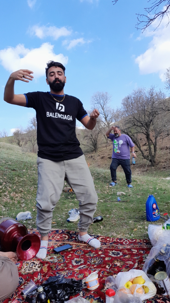 Ein Mann tanzt im Vordergrund, ein anderer im Hintergrund. Auf dem darunter liegenden Teppich auf der Wiese ist das leckere Essen angerichtet.