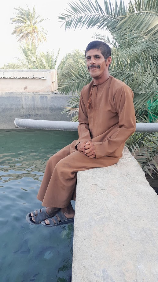 Jamal sitzt auf einer Mauer neben einem Wassertrog zur Linken. Er trägt einen lehmbraunen Kaftan und lacht in die Kamera (uns zu).