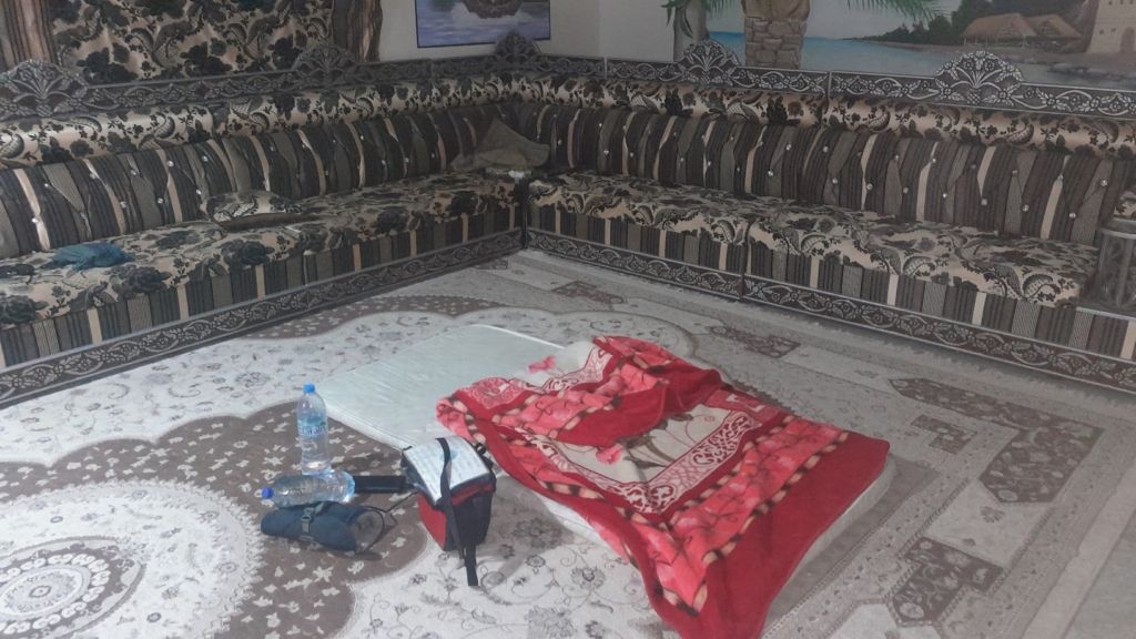 Blick in ein arabisches Zimmer. Der Boden ist mit einem sandbraunen Teppich mit hellbraunem Blumenmuster bedeckt. Braune Sofas mit mattgoldenem Muster stehen an den Wänden und in der Ecke. In der Mitte des Raumes liegt meine improvisierte Schlafstätte: eine dünne weiße Matratze und eine dicke rote Pferdedecke. Daneben stehen zwei Wasserflaschen und meine Tasche für Elektronik.