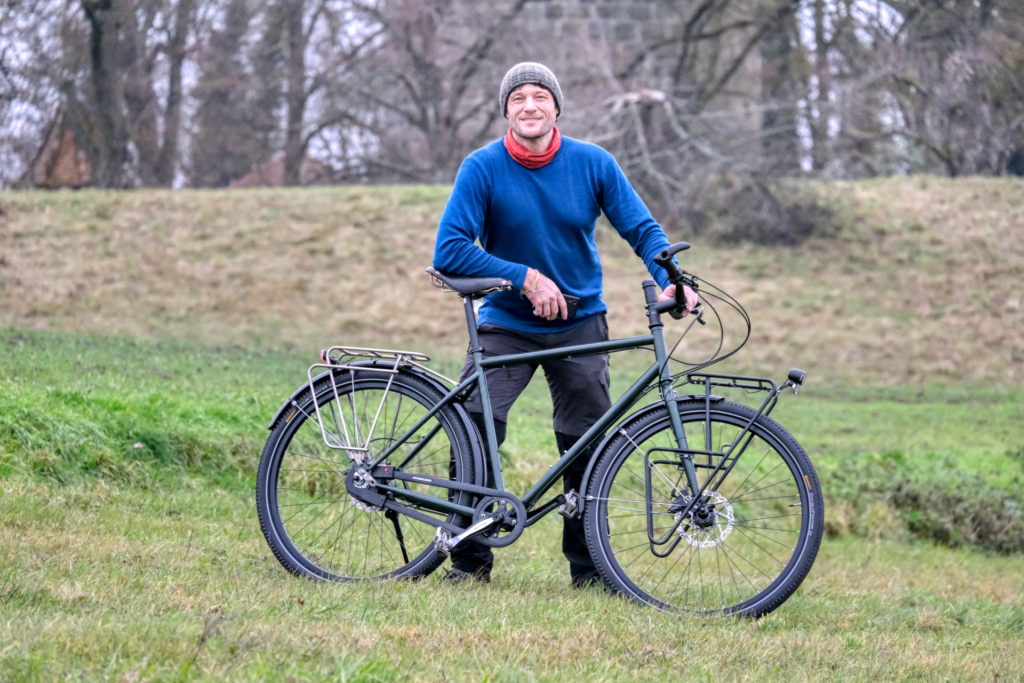 Es ist sichtbar kalt. Ich stehe im Fokus in der Mitte einer wintergrünen Wiese. Ich habe einen leuchtend blauen Pullover an, eine graue Strickmütze und einen roten Schlauchschal. Ich grinse glücklich und lehne entspannt auf einem mattgrünen Reiserad ohne Taschen.