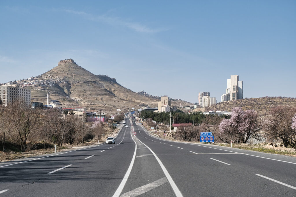 Eine graue Autobahn führt in die Ausläufer von Mardin, links überragt vom beigen Berg mit der Burg auf der steilen Spitze.