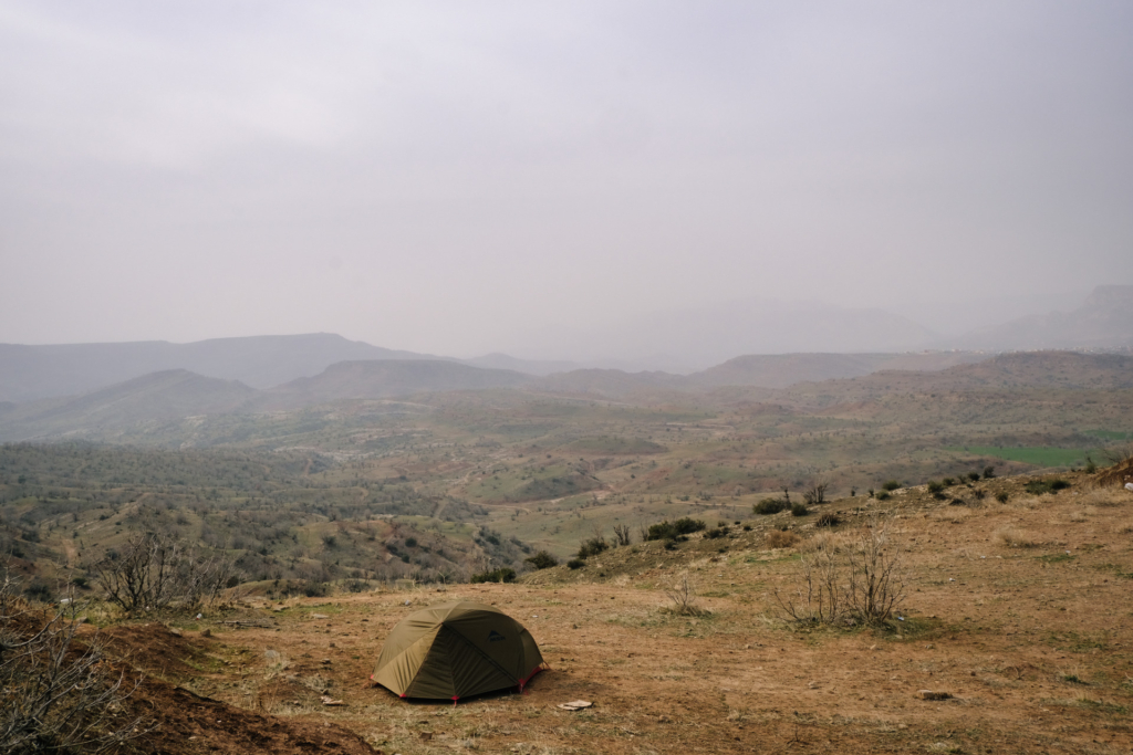 Grünes Zelt auf trockenem Boden vor einer weitläufigen, dunstigen Berglandschaft unter bewölktem Himmel.