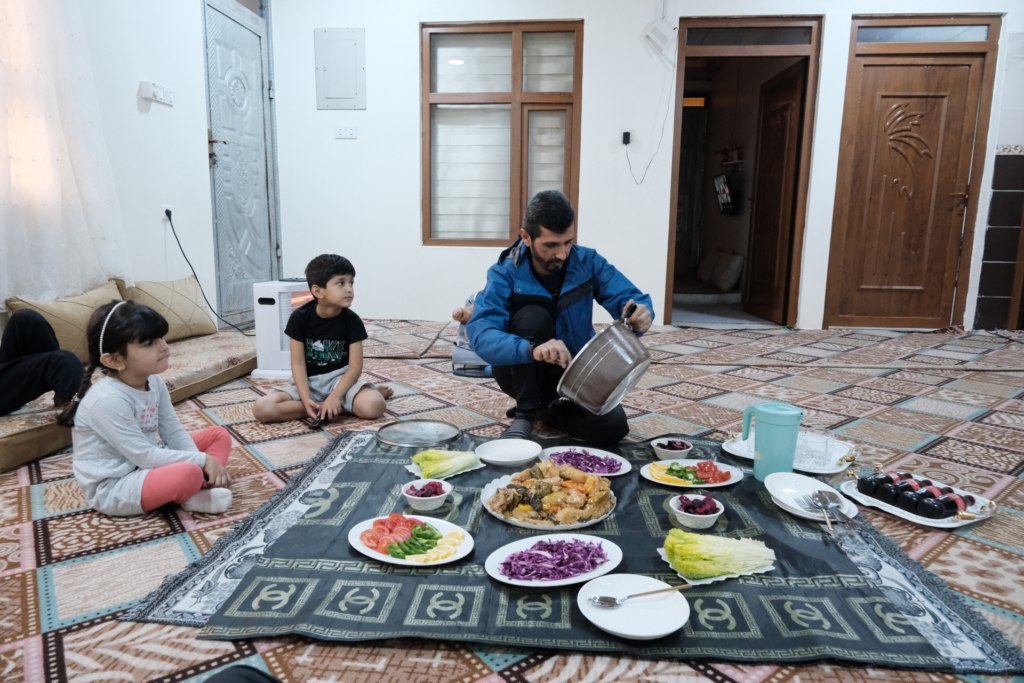 Rahim serviert Essen, das auf dem Boden angerichtet wurde. Mehrere Kinder sitzen gespannt daneben und beobachten die Szene.