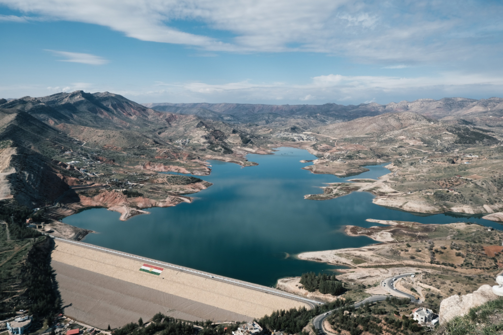 Blick auf den tief türkisen Stausee von Duhok, umgeben von Bergen. Eine Straße schlängelt sich entlang des Sees, im Vordergrund der Damm mit Flagge.