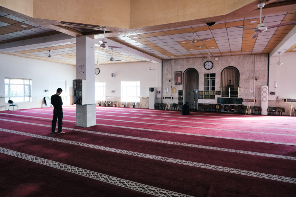 Imam Rahim betet in seiner Moschee, an einer weißen Säule und auf einem rot gestreiften Teppich.