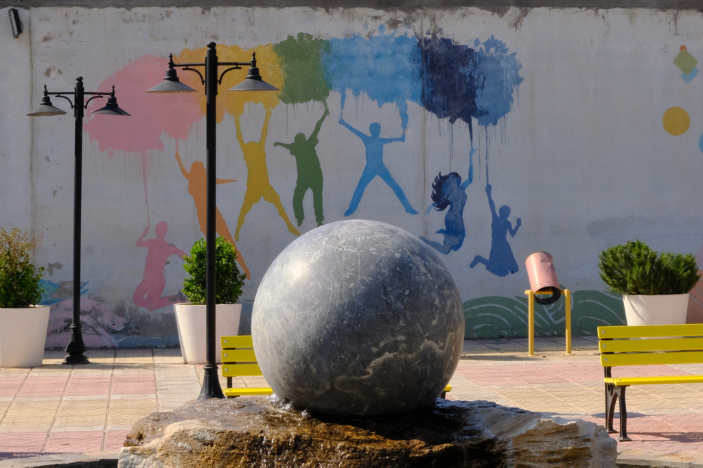 Die Kunstinstallation im Park von Erbil zeigt eine große Marmorkugel auf einem Brunnen. An der Wand dahinter springen abstrakte, regenbogenfarbene Figuren mit Ballons in der Hand über die Kugel; die Frauen sind mit offen wehenden Haaren dargestellt.