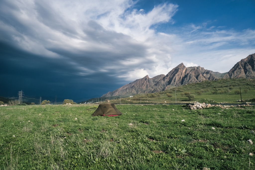 Zelt auf grüner Wiese, umgeben von schroffen, steilen Bergen rechts. Von links zieht ein schweres Gewitter heran.