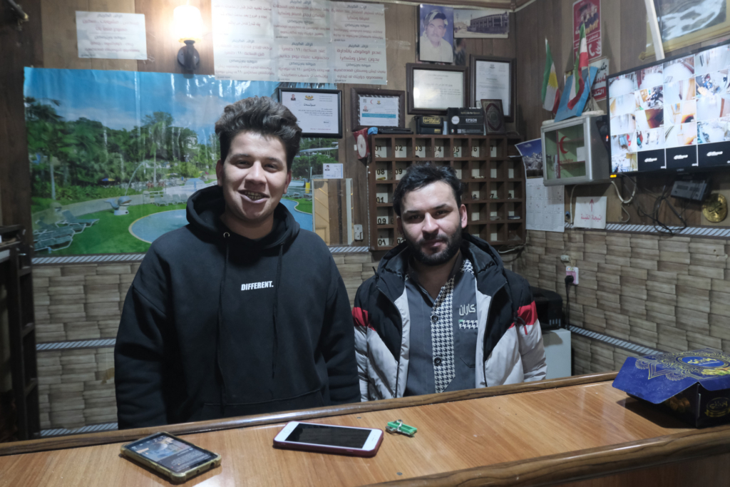 Ein junger Mann und ein Mann mittleren Alters mit Vollbart stehen hinter der Rezeption eines altmodischen Hotels.