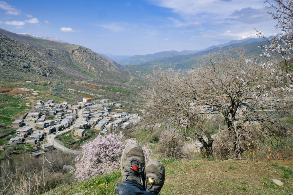 Blick über meine Füße ins bergige Tal, vorbei an rosa Mandelblüten. Auf meinen Schuhen liegen rote und goldene Schokoherzen
