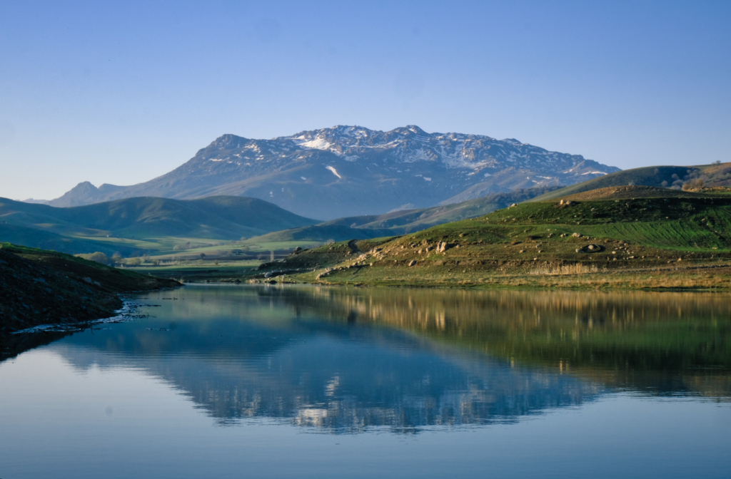Blick über einen stillen Stausee, in dem sich ein graues Bergmassiv spiegelt.
