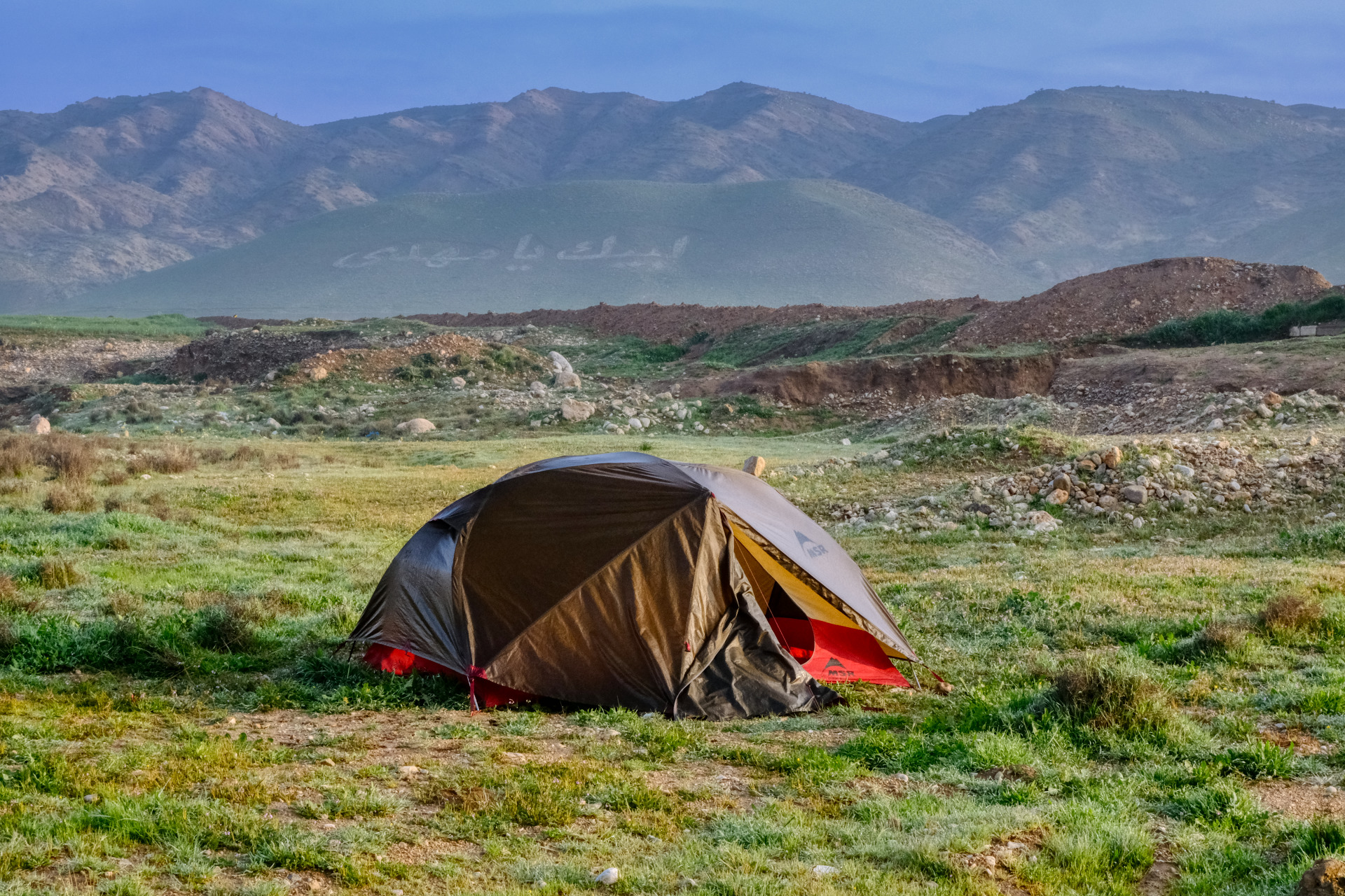 Das Nasse Zelt wird von der prallen Sonne beschienen. Auf dem Berg im Hintergrund steht in kurdischer Schrift "Wir sind ewig"
