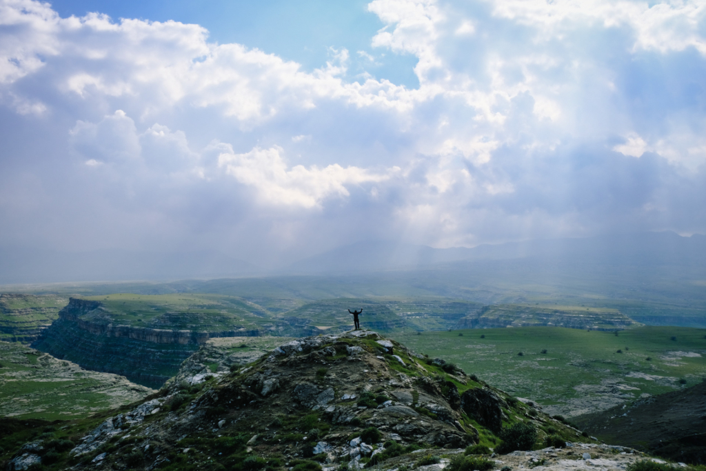 Ich stehe in mittlerer Entfernung auf einer Bergspitze. Hinter mir erstrecken sich massive Schluchten und die Sonne bricht durch den wolkenbehangenen Himmel