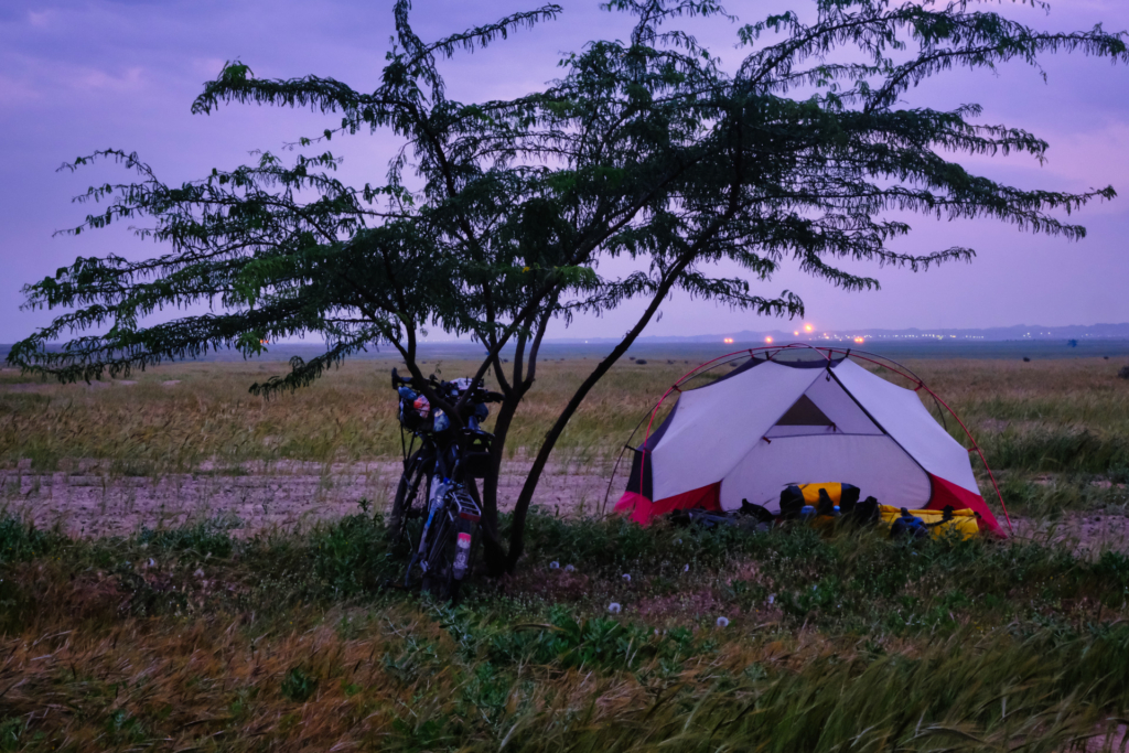 Das Zelt ist unter einem sehr dürren Baum, aufgebaut. Es dämmert schon und im Hintergrund sind die Flammen einer Raffinerie über dem Zelt zu sehen