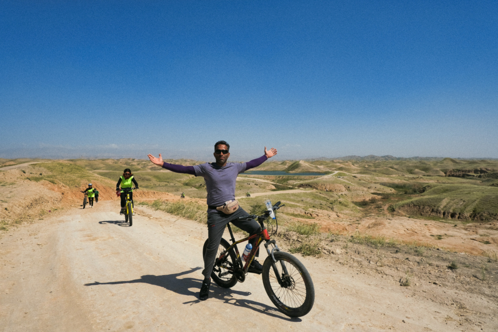 Ein Feldweg führt von einem See im Hintergrund durch die Steppe bis zur Kamera. Direkt in der Front präsentiert sich ein Radfahrer mit ausgestreckten Armen, während der Rest der Gruppe noch den Berg hochtrampelt und schiebt.