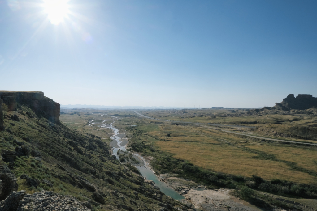 Wir schauen von einem Berg in das grüne Flachland. Zu sehen sind ein Fluss mit wenig Wasser und eine Straße, die bis zum Horizont führt.