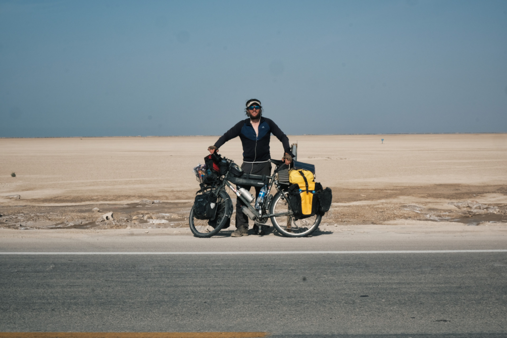 Ich stehe mit dem beladenen Reiserad am Straßenrand mit Blick auf einen endlos tiefen Strand, der wie eine Wüste wirkt. 