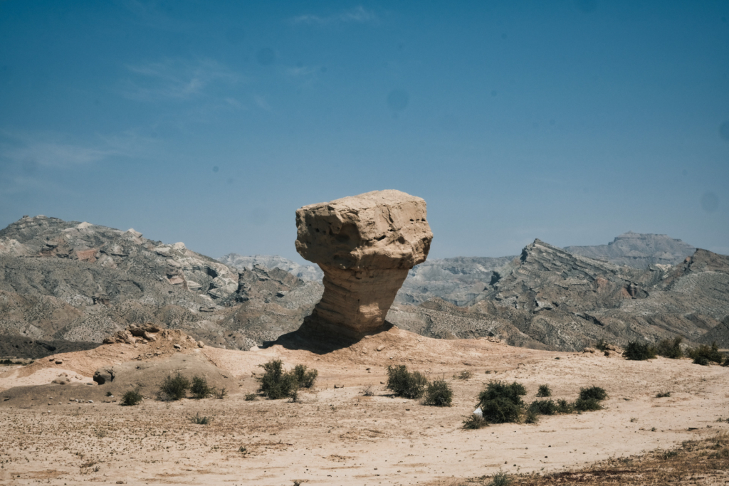 Zentriert im Bild steht ein Felsen, der wie ein Quader auf einer schrägen Säule thront. Die Farben von Boden und Felsen sind sandig. Im Hintergrund sind sehr schroffe, fast außerirdisch wirkende graue Felsformationen zu sehen