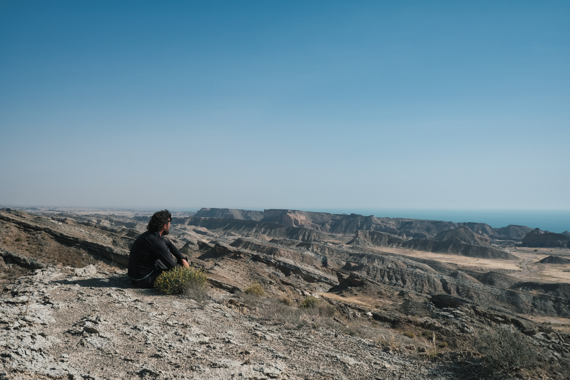 Ich sitze auf einem felsigen, kargen Hügel und blicke über eine weite, trockene, zerklüftete Landschaft aus braun-beigen Felsformationen und Tälern, die bis zum Horizont reichen. Am Horizont ist das blaue Meer unter einem klaren, hellblauen Himmel zu sehen.