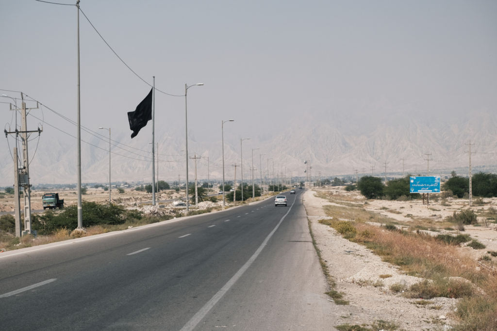 Eine Straße führt geradeaus weiter. Die Berge am Horizont sind vor lauter Smog nicht mehr zu erkennen. Am rechten Straßenrand weht eine schwarze Flagge
