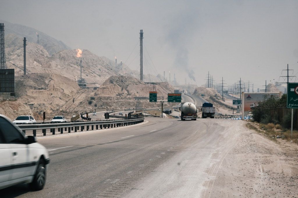 Eine geschwungene Straße führt durch eine smogbelastete, karge, graubraune Hügellandschaft. Überall ragen Türme mit Pilotflammen aus dem Boden. In der Entfernung sind Rauchschlote und auf den Straßen Tanklaster zu sehen.