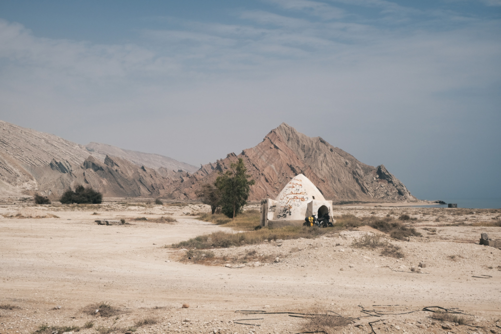 Vor einer weißen Wasserzisterne steht mein Rad. Im Hintergrund steil aufragende Lehmfarbene Berge