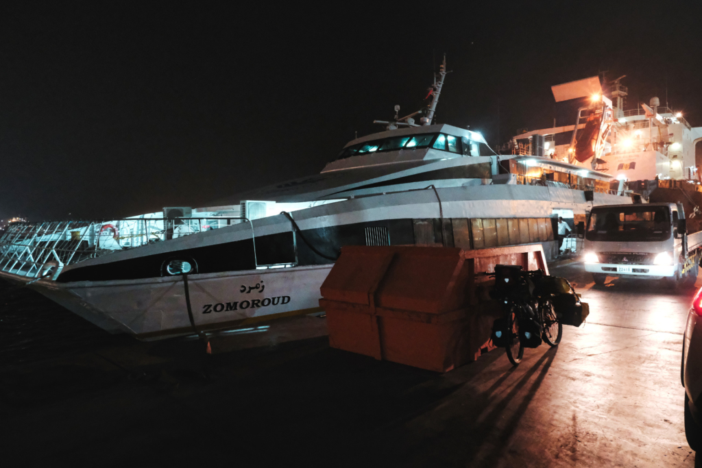 Nachtaufnahme: Im Hintergrund ist eine Fähre zu sehen, die im Dunkeln eher wie eine Yacht wirkt. Im Vordergrund steht mein beladenes Fahrrad, beleuchtet von einem Polizeiauto.