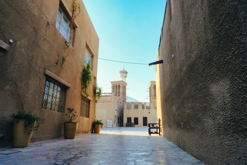 Eine weite, schattige Gasse führt durch eine arabische Altstadt, deren sandfarbene Hauswände links und rechts aufragen. Weiter vorne ist der Beginn eines sonnigen Marktplatzes zu sehen.