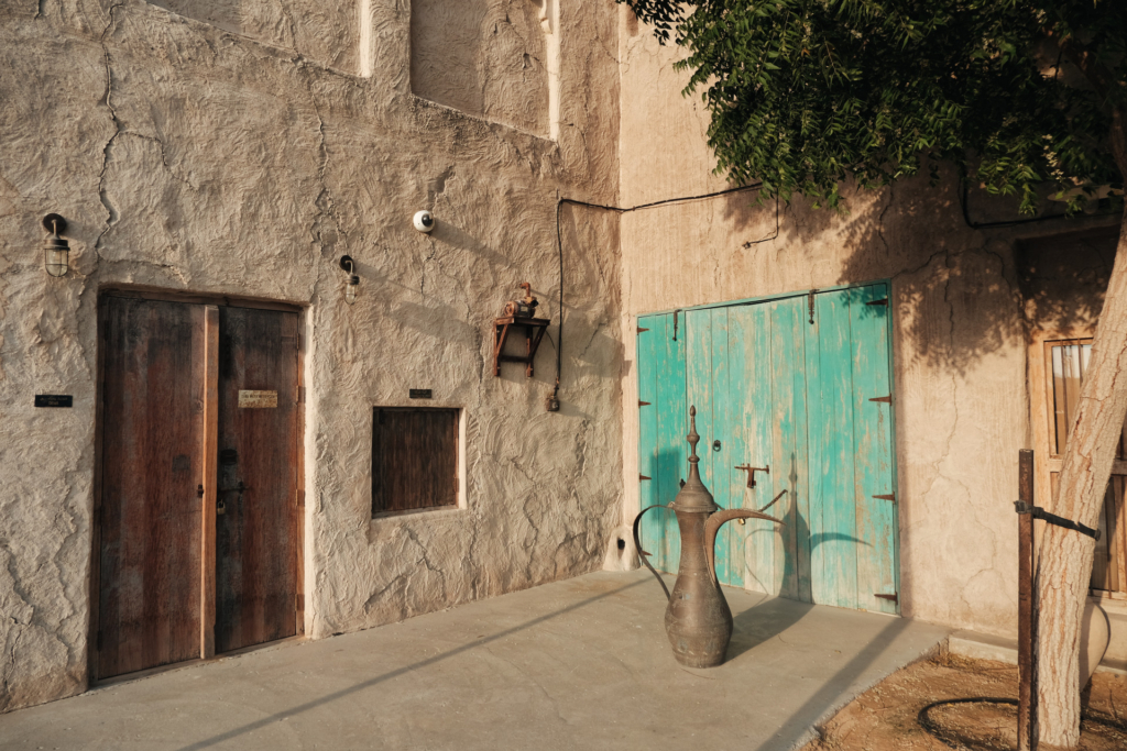 Ein Hauseck im altmodischen arabischen Stil mit Wänden in gedeckten Sandfarben. Links befindet sich eine walnussbraune Doppeltür aus Holz, rechts eine türkise Tür. Vor der türkisen Tür steht eine übergroße arabische Kaffeekanne.
