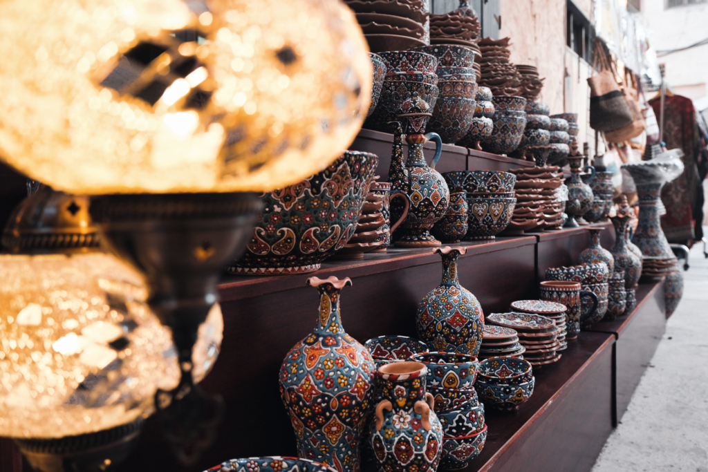 Links im Vordergrund hängen unscharf beleuchtete Lampions. Dahinter erstrecken sich auf drei Regalebenen kunstvoll gefertigte Töpferwaren