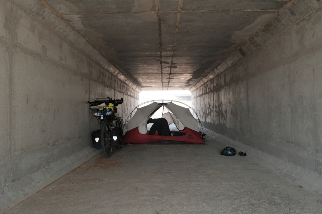 Ein niedriger Tunnel, dessen Licht am vorderen Ende leicht blendet. Links lehnt das Fahrrad an der Wand. In der Mitte steht das Innenzelt quer zum Tunnel, und ich liege darin.