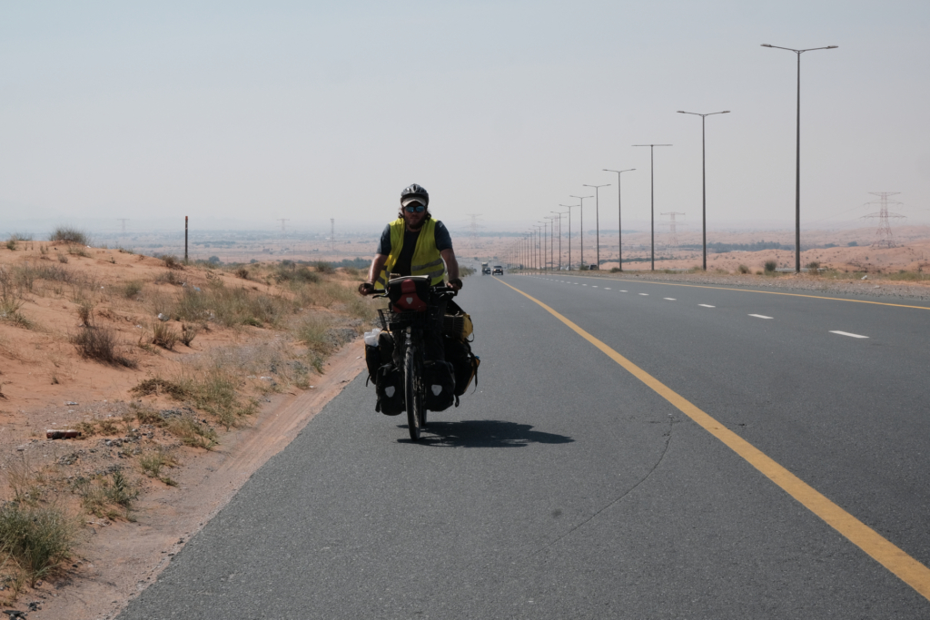 Ich fahre auf dem Seitenstreifen, mit dem Fahrrad auf die Kamera zu. Links und Rechts von der Straße ist die Sandwüste zu sehen und das Wetter ist sehr dunstig