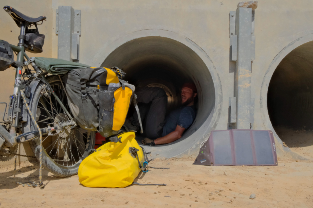 Eine Betonröhre in der Mitte des Bildes. Ich liege erschöpft darin, die Beine an die eine Wand und den Rücken an die andere gekrümmt. Meine Zunge hängt aufgrund der Hitze aus dem Mund. Neben der Röhre steht das noch voll bepackte Fahrrad.