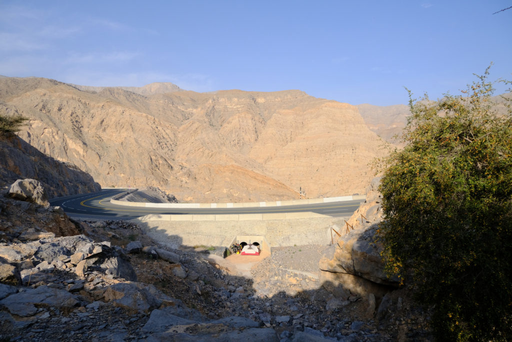Eine Straße führt erhöht über eine Schlucht durch die sandfarbenen Berge. Am Fuße der Straße steht mein Zelt mitten in der Sonne