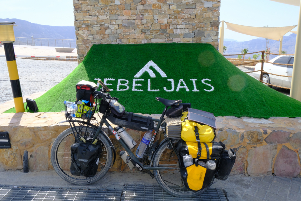 Das beladene Fahrrad lehnt an einer Mauer. Hinter der Mauer steht in weißer Schrift auf grünem Kunstrasen: Jebel Jais.