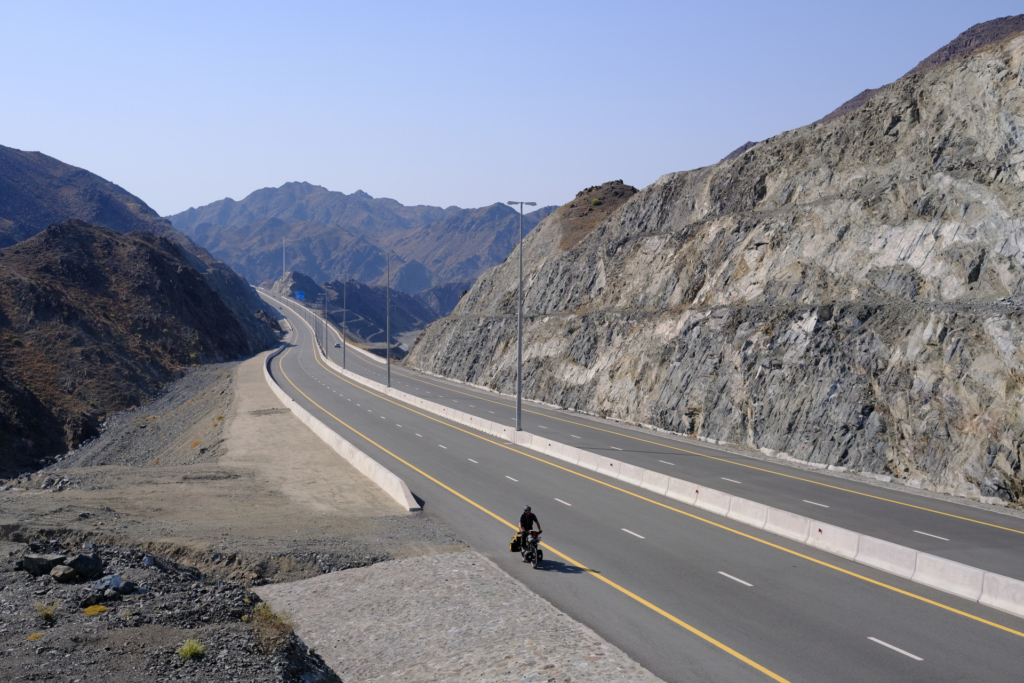 Eine Straße führt durch die Berge auf uns zu. Wir schauen von oben darauf und sehen mich mit dem Fahrrad entlang fahren