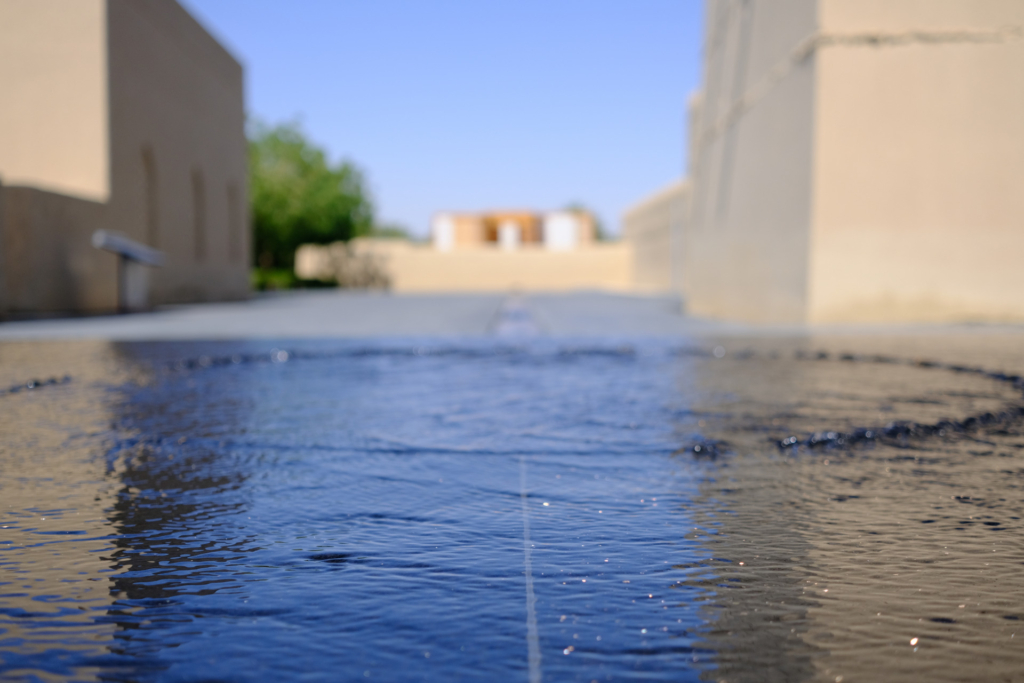 Die komplette untere Bildhälfte besteht aus einer flachen Wasserfläche auf dunklem Stein. Ein konzentrischer Ring von Wellen geht von der Mitte aus. Das Bild ist komplett auf das Wasser fokussiert. Im Hintergrund sind links und rechts stark verschwommene, sandfarbene Steinmauern zu erkennen.