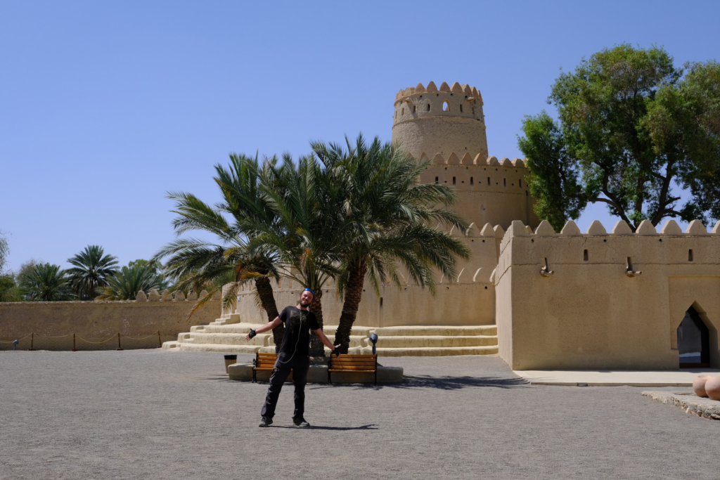 Ich stehe mittig im Bild im Al Jahil Fort. Die Sonne brennt herunter. Direkt hinter mir sieht man eine dreistämmige Palme. Rechs dahinter ist der runde Wachturm des Forts zu sehen.